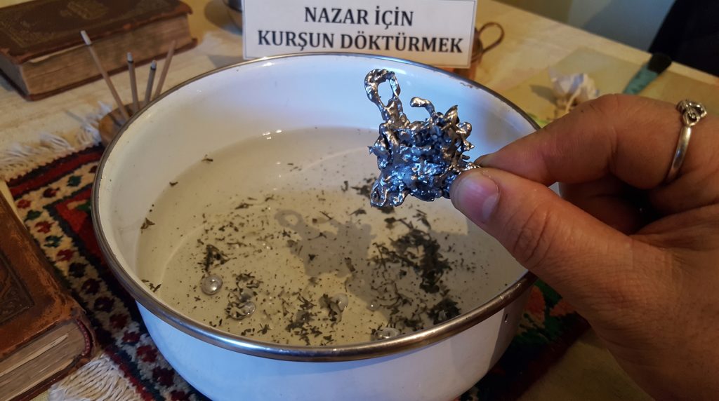Kurşun dökme ritüelinden bir an: Sağ tarafta bir el, su dolu beyaz emaye kase üzerinde soğuyup şekil almış parlak metalik kurşun parçasını tutuyor. Arka planda üst kısımda "NAZAR İÇİN KURŞUN DÖKTÜRMEK" yazılı bir tabela yer alıyor.